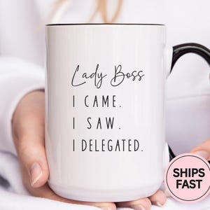 May include: White ceramic mug with black handle and rim. The mug features the text "Lady Boss" in a script font, followed by "I CAME. I SAW. I DELEGATED." in a simple sans-serif font. A pink and white sticker that says "SHIPS FAST" is on the mug.