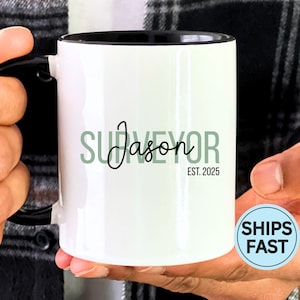 May include: White ceramic mug with a black interior and handle. The mug features the name "Jason" in cursive, with the word "SURVEYOR" above and "EST. 2025" below, all in a green font. A "SHIPS FAST" sticker is visible.