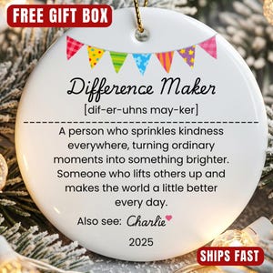 以下が含まれることがあります： 「Difference Maker」の文字と定義が書かれた白いセラミック製のクリスマスオーナメント。カラフルなバナーと「Also see: Charlie」の文字、2025年が装飾されています。