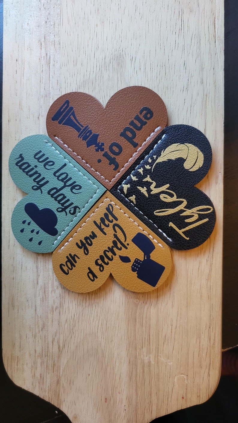May include: Four heart-shaped leather bookmarks with different designs. One bookmark features a chess piece, another has the text "we love rainy days", another has the text "can you keep a secret?" and the last one has the text "forever" with a feather.