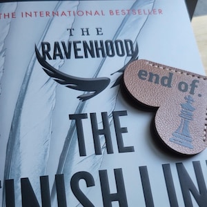 May include: A brown leather heart-shaped bookmark with the text "end of." and a black chess piece illustration. The bookmark is on top of a book titled "The Ravenhood The Finish Line" with the text "The International Bestseller" at the top.