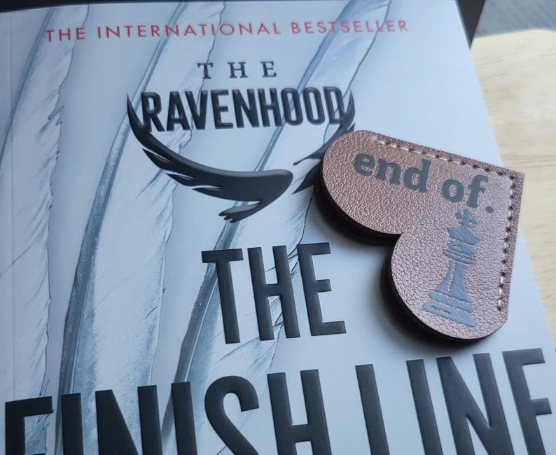 May include: A brown leather heart-shaped bookmark with the text "end of." and a black chess piece illustration. The bookmark is on top of a book titled "The Ravenhood The Finish Line" with the text "The International Bestseller" at the top.