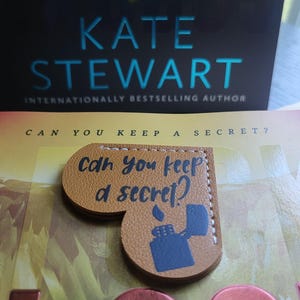 May include: A brown leather heart-shaped bookmark with the text "Can you keep a secret?" and a black silhouette of a lighter. The bookmark is on top of a book with the title "Can You Keep A Secret?" by Kate Stewart, an internationally bestselling author.