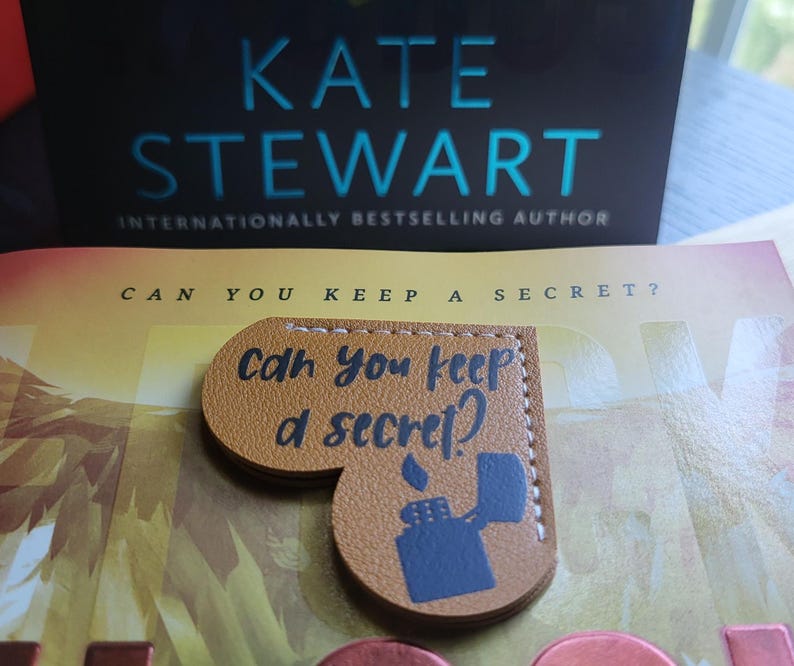 May include: A brown leather heart-shaped bookmark with the text "Can you keep a secret?" and a black silhouette of a lighter. The bookmark is on top of a book with the title "Can You Keep A Secret?" by Kate Stewart, an internationally bestselling author.