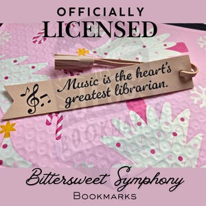 Peut inclure: Un marque-page rose clair avec le texte "Music is the heart's greatest librarian." présente une clé de sol et des notes de musique. Le marque-page a un pompon et une boucle. Le fond est un papier bulle rose avec des motifs de sapins de Noël et de flocons de neige. Le texte "OFFICIALLY LICENSED" et "Bittersweet Symphony Bookmarks" sont également visibles.