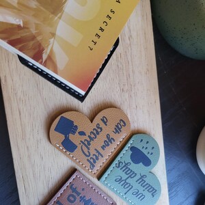May include: Three heart-shaped leather bookmarks with different designs. The first bookmark is brown with a chess piece and the text "end of". The second bookmark is green with a cloud and raindrops and the text "rainy days we love shop". The third bookmark is brown with a teacup and the text "Can you keep a secret".
