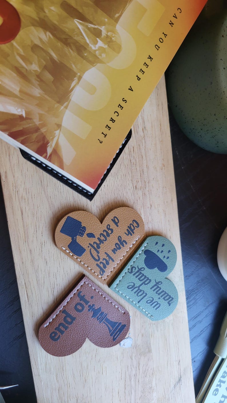 May include: Three heart-shaped leather bookmarks with different designs. The first bookmark is brown with a chess piece and the text "end of". The second bookmark is green with a cloud and raindrops and the text "rainy days we love shop". The third bookmark is brown with a teacup and the text "Can you keep a secret".