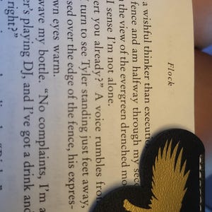 May include: A close-up of a black leather bookmark with a gold eagle design. The bookmark is folded in half and is resting on a page of a book. The text on the page reads "More of a wishful thinker than executioner. Against the fence and am halfway through my second cider, lost in the view of the evergreen drenched mountains when I sense I'm not alone. "Sean desert you already?" A voice rumbles from next to me. I turn to see Tyler standing just feet away, his arms crossed over the edge of the fence, his expression and brown eyes warm. "Yeah," I wave my bottle. "No complaints, I'm a fan of whoever's playing DJ, and I've got a drink and a view. Tyler, right?"
