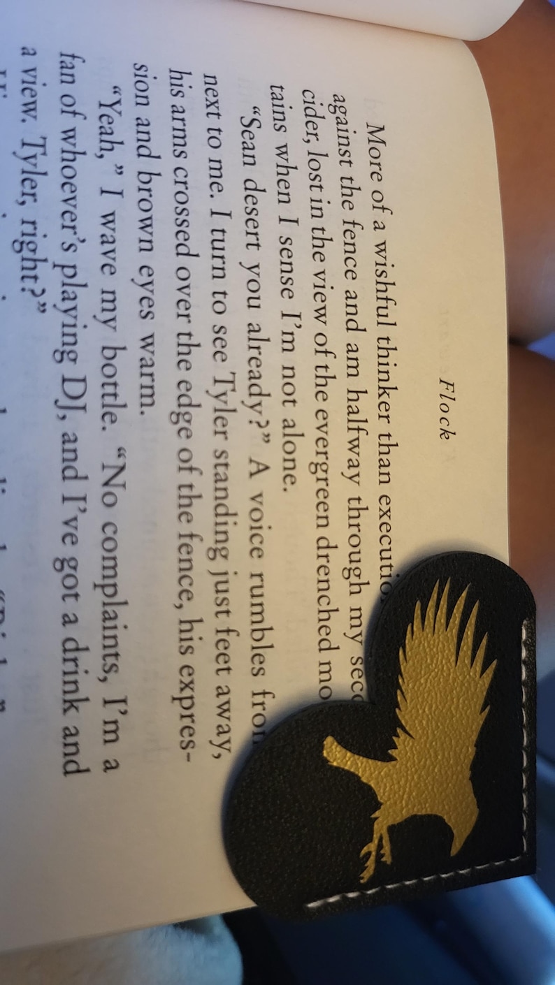 May include: A close-up of a black leather bookmark with a gold eagle design. The bookmark is folded in half and is resting on a page of a book. The text on the page reads "More of a wishful thinker than executioner. Against the fence and am halfway through my second cider, lost in the view of the evergreen drenched mountains when I sense I'm not alone. "Sean desert you already?" A voice rumbles from next to me. I turn to see Tyler standing just feet away, his arms crossed over the edge of the fence, his expression and brown eyes warm. "Yeah," I wave my bottle. "No complaints, I'm a fan of whoever's playing DJ, and I've got a drink and a view. Tyler, right?"