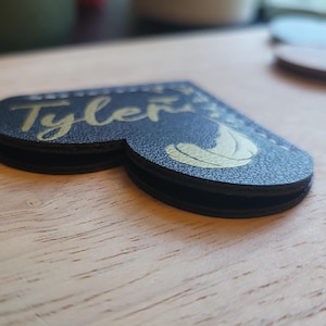 May include: A black leather heart-shaped bookmark with the name "Tyler" written in gold lettering. A gold feather design is also visible on the bookmark.