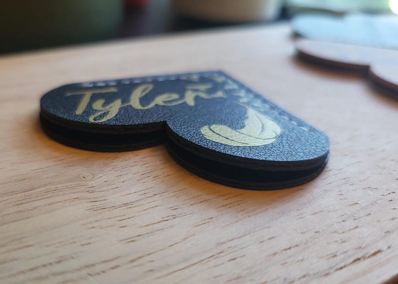 May include: A black leather heart-shaped bookmark with the name "Tyler" written in gold lettering. A gold feather design is also visible on the bookmark.