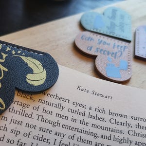 May include: Three heart-shaped leather bookmarks with different designs and text. The first bookmark is black with gold lettering and a feather design. The text reads "Tyler". The second bookmark is light blue with white lettering and a design of a watering can. The text reads "Can you keep a secret?" The third bookmark is brown with white lettering and a design of a bird. The text reads "Jo pua".
