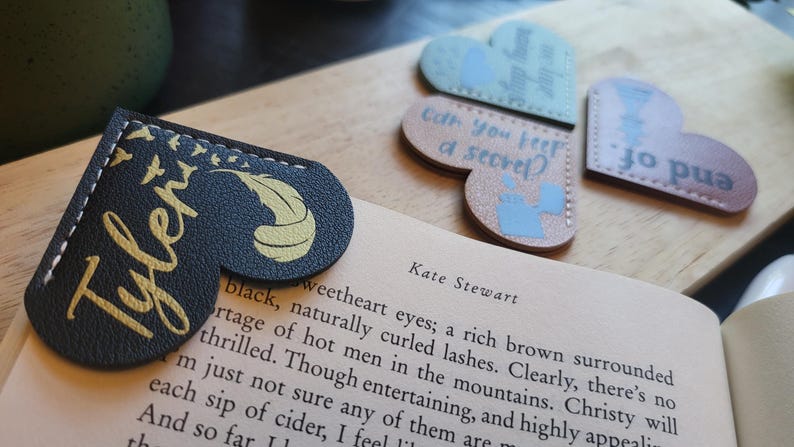 May include: Three heart-shaped leather bookmarks with different designs and text. The first bookmark is black with gold lettering and a feather design. The text reads "Tyler". The second bookmark is light blue with white lettering and a design of a watering can. The text reads "Can you keep a secret?" The third bookmark is brown with white lettering and a design of a bird. The text reads "Jo pua".