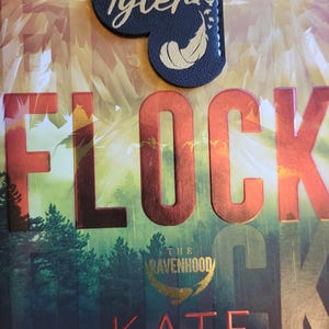 May include: A book titled "Flock: The Ravenhood" by Kate Stewart, an internationally bestselling author. The book cover features a red and gold title with a forest background. A black leather bookmark with a gold feather and the word "Tyler" is on the book.