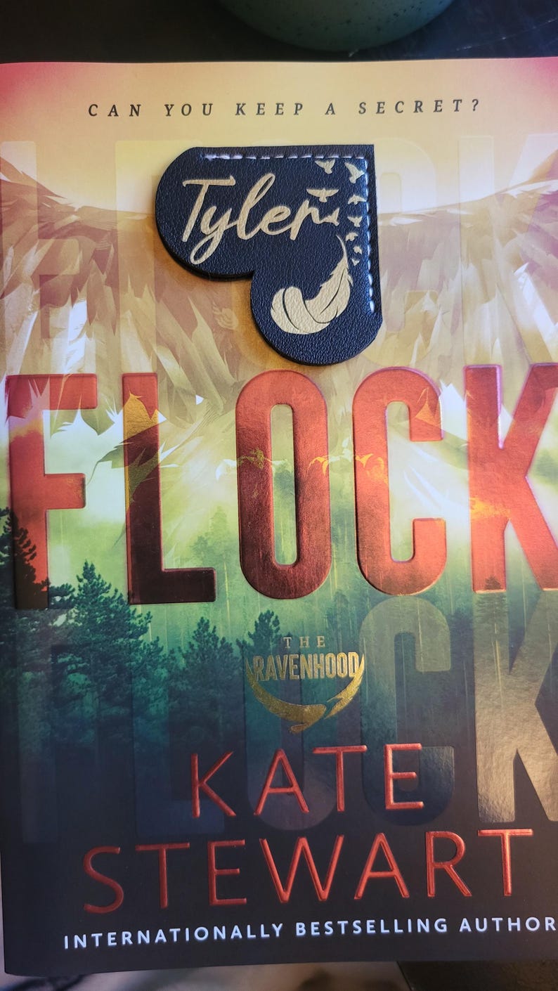 May include: A book titled "Flock: The Ravenhood" by Kate Stewart, an internationally bestselling author. The book cover features a red and gold title with a forest background. A black leather bookmark with a gold feather and the word "Tyler" is on the book.