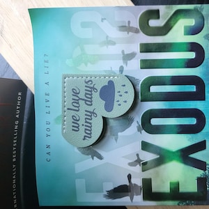 May include: A green heart-shaped bookmark with the text "we love rainy days" and a cloud with raindrops. The bookmark is on top of a book with the title "Exodus" in large, black letters.