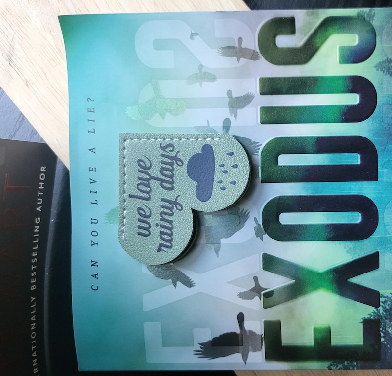 May include: A green heart-shaped bookmark with the text "we love rainy days" and a cloud with raindrops. The bookmark is on top of a book with the title "Exodus" in large, black letters.