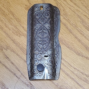 May include: A rectangular, dark metallic plate with a curved profile and intricate, embossed scrollwork patterns. The piece features a small circular hole at the top, a central horizontal slot, and a small, dark circular button near the bottom edge.