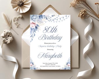 May include: An invitation for an 80th birthday party. The invitation is white with blue and pink floral accents. The text reads "80th Birthday" and "Marybeth". The invitation is inside a beige envelope, with white ribbon and decorative elements.