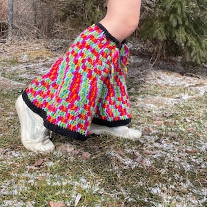 Puede incluir: Un calienta piernas de crochet colorido con un borde negro. El calienta piernas está hecho con una variedad de colores brillantes, incluyendo rosa, rojo, naranja, amarillo, verde y azul.