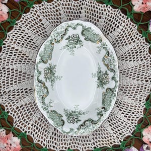 May include: An oval white ceramic serving platter featuring a decorative border with green floral patterns, intricate scrollwork, and gold-toned accents. The center of the platter is plain white, surrounded by a detailed rim design.