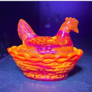 Könnte beinhalten: Ein leuchtend orange-rosa Glas Henne-auf-Nest-Schale. Die Henne sitzt auf einem Korbgeflecht-Sockel, mit detailliertem Schwanz und Kopf. Das Glas hat eine glänzende Oberfläche, die das Licht reflektiert. Ein dekorativer Küchenartikel.