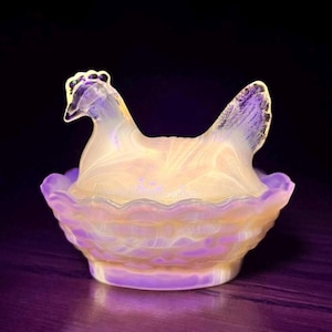May include: A decorative glass hen-shaped dish, with a ruffled edge, in shades of yellow, white, and purple. The hen sits in a nest-like bowl. The glass has a marbled appearance, and the background is dark.