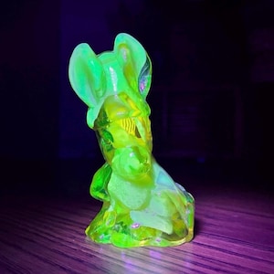 May include: A vibrant, glowing green glass sculpture of a rabbit-like figure, with prominent ears and a rounded form. The figurine has a textured surface and is lit with a bright, neon-like glow, set against a dark backdrop.