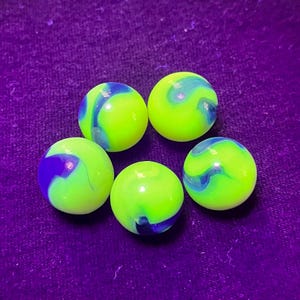 May include: Five marbles with a neon green base color and swirling blue and purple patterns. The marbles are round and smooth, set against a purple background.