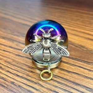 May include: A spherical ornament featuring an iridescent, multi-colored glass orb with a metallic finish. A silver-toned metal bee figurine with detailed wings and body is attached to the front. A small gold-toned metal ring hangs from the base.