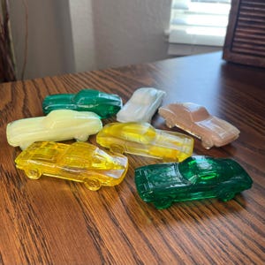 May include: Seven small, transparent toy cars in shades of yellow, green, and white are displayed. The toy cars are arranged on a wooden surface, showcasing their classic car design.