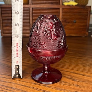 Pode incluir: Um castiçal de vidro vermelho rubi em forma de ovo com detalhes florais e de videira. O castiçal tem aproximadamente 10 cm de altura. A base é um pequeno pedestal. A parte superior em forma de ovo tem uma superfície texturizada e um desenho floral.
