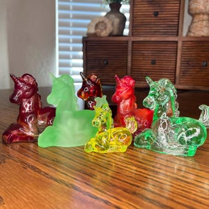 Pode incluir: Uma coleção de figuras de unicórnios de vidro coloridas em tons de vermelho, verde e amarelo. As figuras são exibidas em uma superfície de madeira, com uma janela e itens decorativos ao fundo. Cada unicórnio tem uma crina e um chifre detalhados.