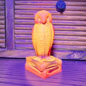 Puede incluir: Una figura de cristal naranja translúcido de un búho posado sobre una pila de dos libros. El búho presenta texturas de plumas detalladas y grandes ojos circulares, mientras que la base consta de dos volúmenes rectangulares con detalles.