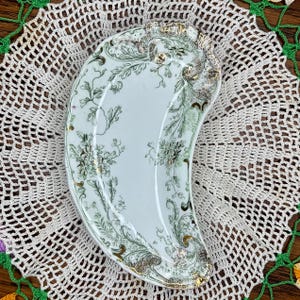 May include: A crescent-shaped ceramic dish featuring a white base with intricate green floral and leafy scrollwork patterns. The edges of the dish are accented with gold trim, and the surface displays a glossy finish with detailed botanical motifs.