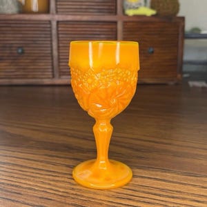 Könnte beinhalten: Ein orangefarbener Glasbecher im Vintage-Stil mit einem detaillierten Blumenmuster auf der Schale. Der Stiel und der Fuß sind ebenfalls orange. Der Becher besteht aus Glas und wird wahrscheinlich zum Servieren von Getränken verwendet. Das Glas steht auf einer Holzoberfläche.