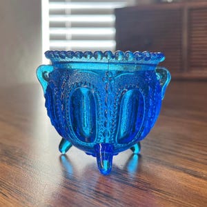 Puede incluir: Un pequeño cuenco de vidrio azul cobalto con un borde festoneado y tres patas. El cuenco tiene un patrón decorativo en relieve.