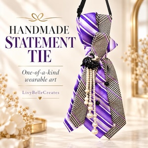 May include: A necktie featuring a mix of purple and white diagonal stripes and a black and white houndstooth pattern. The tie is adorned with a black floral embellishment and multiple strands of white pearls. Text reads: HANDMADE STATEMENT TIE, One-of-a-kind wearable art, LivyBelleCreates.