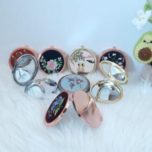 Puede incluir: Una colección de espejos compactos redondos, abiertos y cerrados, con varios diseños, incluyendo bordados florales y un acabado en oro rosa. Los espejos están sobre una superficie blanca y esponjosa.