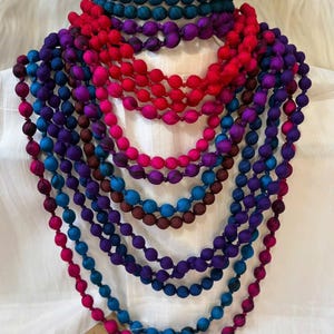 Könnte beinhalten: Eine Sammlung farbenfroher Perlenketten in den Farbtönen Türkis, Rot, Pink, Lila und Blau. Die Halsketten werden auf einer weißen Oberfläche präsentiert, die die Vielfalt der Farben und Designs zeigt. Die Perlen scheinen mit Stoff bedeckt zu sein.
