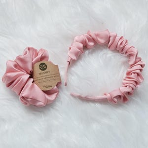 May include: A set of two pink satin hair accessories. One is a scrunchie and the other is a headband. The headband is made of gathered satin fabric. The scrunchie is handmade by Vietnamese artisans. The packaging includes the text "KBT CRAFT HANDWOVEN BY VIETNAMESE ARTISAN Handmade in Vietnam"