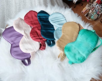 May include: A set of seven silk sleep masks in various colors, including purple, pink, red, teal, blue, gold, and green. The masks are arranged in a row on a white fluffy surface.