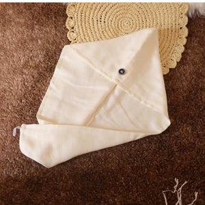 May include: A white cotton hair towel with a button closure. The towel is folded in a triangular shape and is lying on a brown fluffy surface.