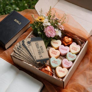 May include: A cardboard gift box containing a bouquet of pink, white, and yellow flowers, a stack of brown tags labeled 50 BIBLE VERSES TO KNOW, and heart-shaped candies with text including HOPE, FAITH, GOD'S WORD, SOLI DEO GLORIA, PRAY ALWAYS, JESUS SAVES, and LOVE.