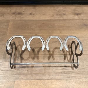 Op de afbeelding: Een metalen schoenenrek gemaakt van paardenhoefijzers. Het rek heeft vijf paardenhoefijzers en een metalen stang om schoenen aan te hangen.