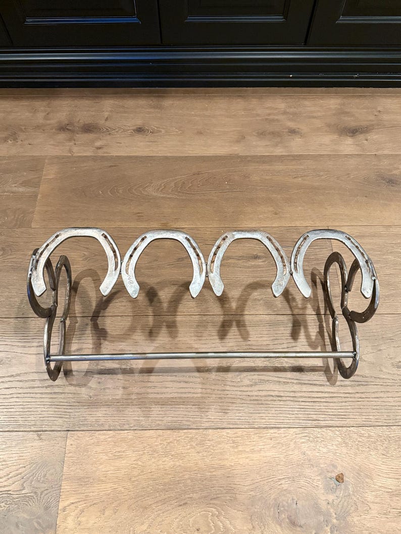 Op de afbeelding: Een metalen schoenenrek gemaakt van paardenhoefijzers. Het rek heeft vijf paardenhoefijzers en een metalen stang om schoenen aan te hangen.