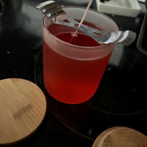 Puede incluir: Una vela roja en un tarro de vidrio transparente con un soporte de mecha de metal. La vela está sobre una superficie negra con una tapa de madera al lado.