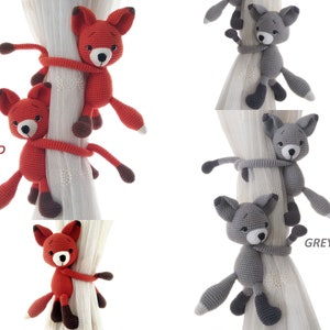 May include: Two crocheted fox curtain tiebacks, one red and one grey, are shown wrapped around a white curtain rod. The tiebacks are made of yarn and have a cute, cartoonish appearance.