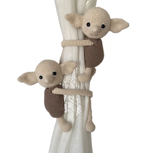 May include: Two crocheted elf dolls in brown and beige colors holding onto a white curtain. The dolls are holding onto the curtain with their arms and legs.