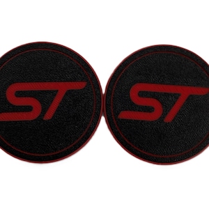 May include: Two black round car coasters with a red "ST" logo. The coasters have a textured surface and a red border. The design is simple and modern, suitable for car interiors.
